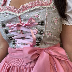 May include: A grey dirndl dress featuring a pink floral pattern, a square neckline with white lace trim, and a pink satin ribbon lace-up bodice with decorative metal eyelets. The dress includes a wide pink satin waist sash tied in a bow.