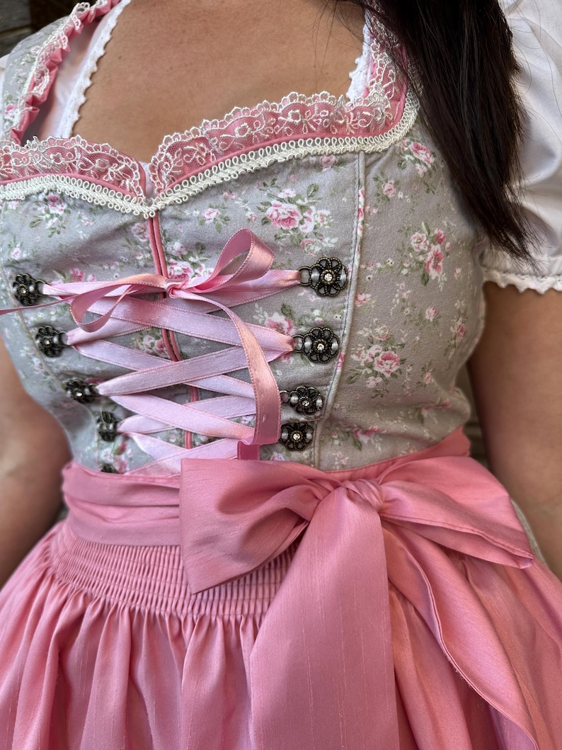 May include: A grey dirndl dress featuring a pink floral pattern, a square neckline with white lace trim, and a pink satin ribbon lace-up bodice with decorative metal eyelets. The dress includes a wide pink satin waist sash tied in a bow.