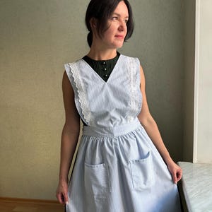 Puede incluir: Un delantal a rayas azules y blancas con ribete de encaje blanco y dos bolsillos delanteros. El delantal es sin mangas y tiene un escote en V. La modelo lleva un top negro debajo del delantal.