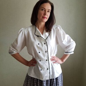 May include: White button-up blouse with black trim and buttons. The blouse has a collar and long sleeves with puffed shoulders.