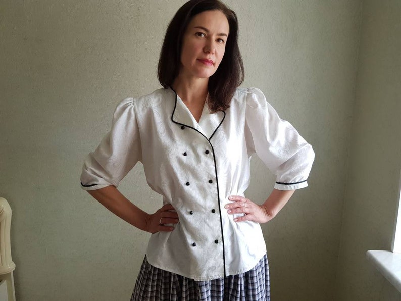 May include: White button-up blouse with black trim and buttons. The blouse has a collar and long sleeves with puffed shoulders.