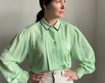 May include: A light green long-sleeved blouse with a white collar and black trim. The blouse has a button-up front and a gathered detail at the shoulders.