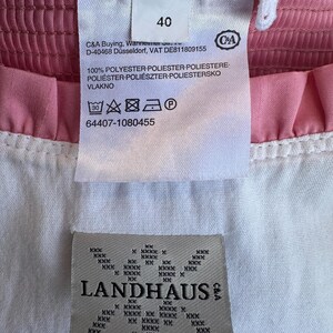 May include: A pink and white garment featuring a white fabric care label with the number 40, C&A branding, and washing instructions. Below it, a beige label displays the text LANDHAUS with a repeating cross-stitch pattern design.