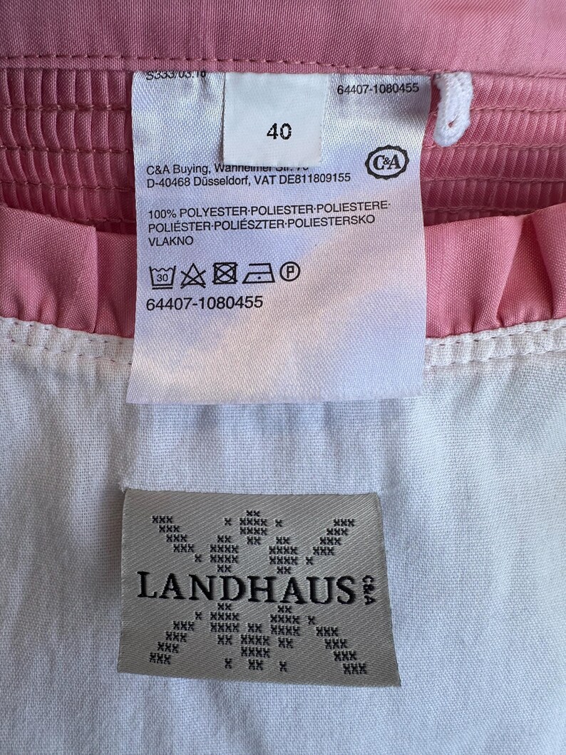 May include: A pink and white garment featuring a white fabric care label with the number 40, C&A branding, and washing instructions. Below it, a beige label displays the text LANDHAUS with a repeating cross-stitch pattern design.