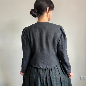 Puede incluir: Una chaqueta de lino negra con mangas largas y hombros ligeramente abullonados. La chaqueta tiene cuello en V y una longitud recortada. La modelo lleva la chaqueta sobre una falda verde oscuro con estampado floral.