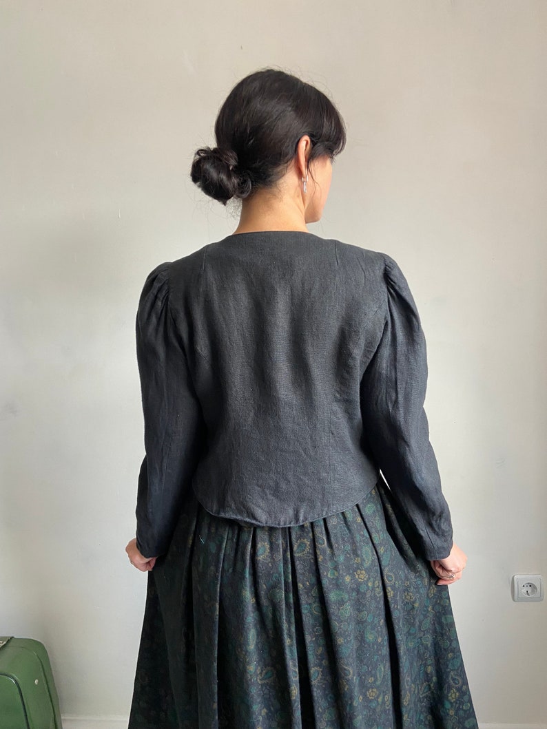 Puede incluir: Una chaqueta de lino negra con mangas largas y hombros ligeramente abullonados. La chaqueta tiene cuello en V y una longitud recortada. La modelo lleva la chaqueta sobre una falda verde oscuro con estampado floral.
