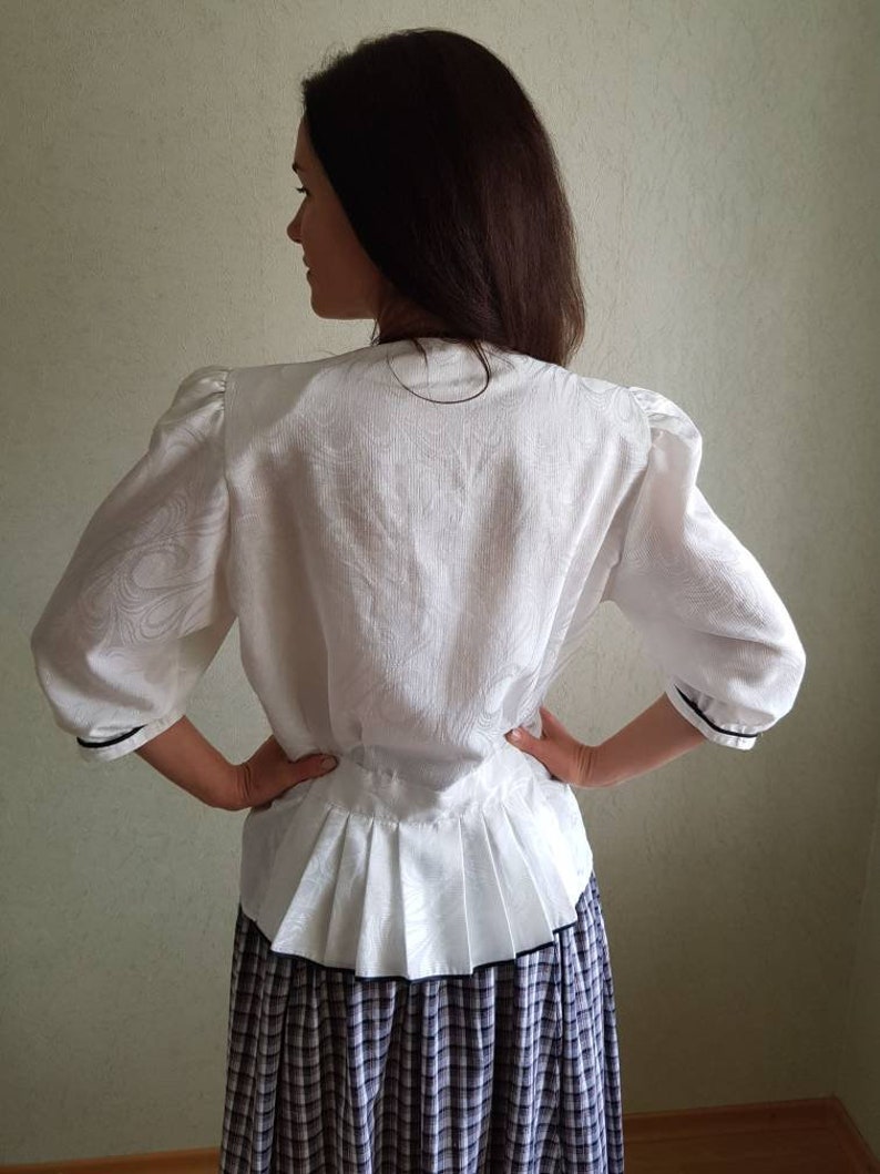 May include: White blouse with a ruffled hem and black trim. The blouse has a fitted bodice and 3/4 sleeves. The model is wearing a plaid skirt.