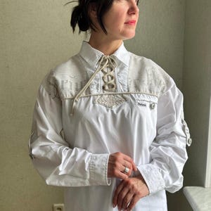 Pode incluir: Camisa branca de manga comprida com colar de renda e detalhes bordados. A camisa tem um logotipo "Schuhplattler" no peito esquerdo.
