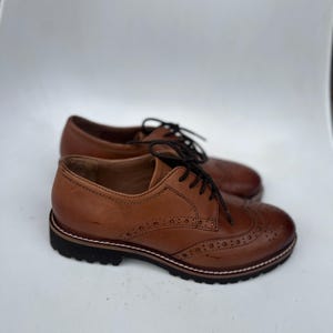 May include: A pair of brown leather Oxford shoes with black laces. The shoes have a classic design with decorative perforations on the toe and sides. The soles are black with a white stitch detail.
