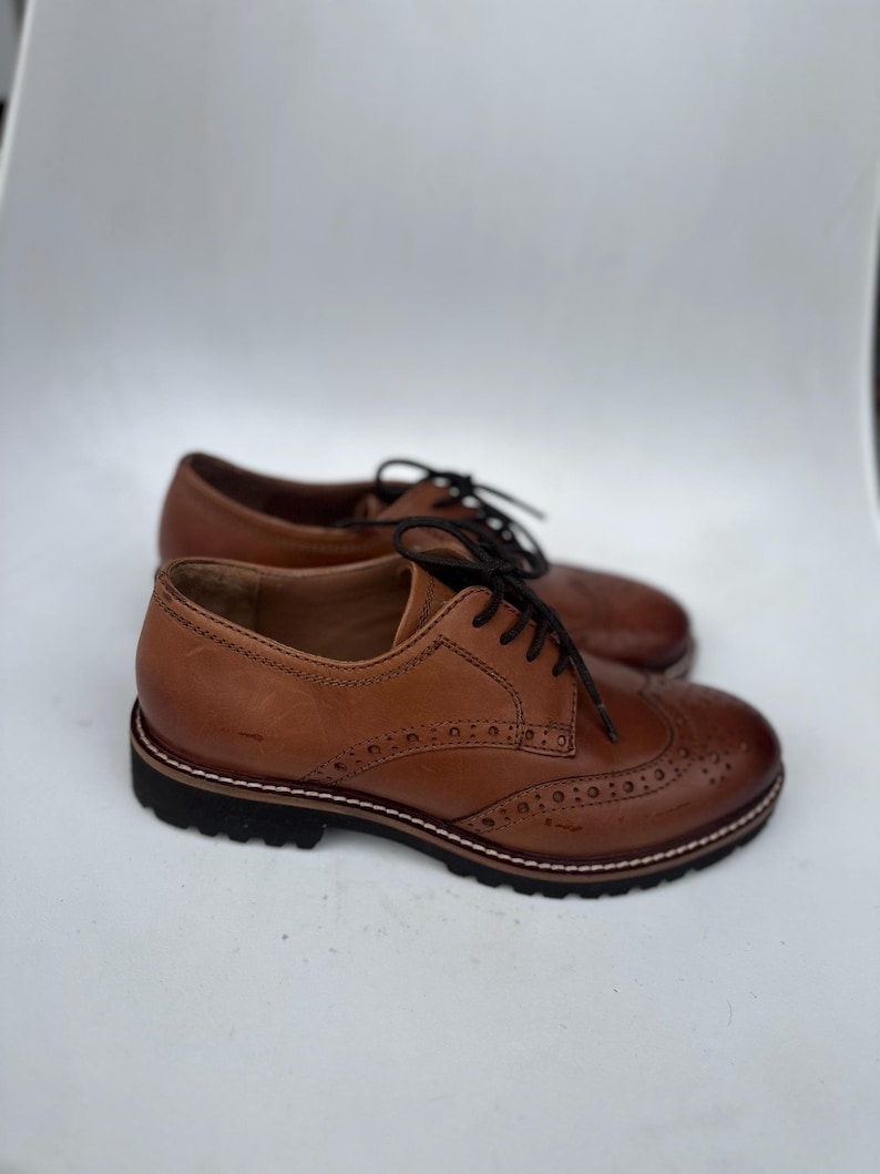 May include: A pair of brown leather Oxford shoes with black laces. The shoes have a classic design with decorative perforations on the toe and sides. The soles are black with a white stitch detail.