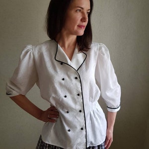 May include: A white button-up blouse with black trim and buttons. The blouse has a slightly puffed sleeve and a fitted waist.