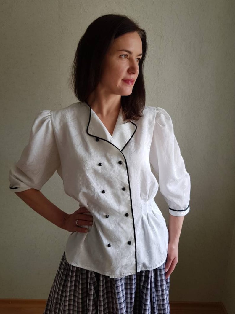 May include: A white button-up blouse with black trim and buttons. The blouse has a slightly puffed sleeve and a fitted waist.