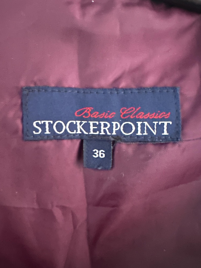 Puede incluir: Un interior de prenda de color morado oscuro con una etiqueta de tela azul rectangular que presenta el texto Basic Classics STOCKERPOINT en letras blancas y rojas. Una etiqueta azul m&aacute;s peque&ntilde;a debajo muestra el n&uacute;mero 36 en blanco.