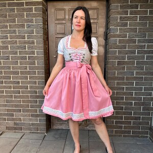 May include: A dirndl dress featuring a white blouse with short puff sleeves, a grey bodice with pink lace-up detailing and metal hooks, and a solid pink skirt with a wide matching waist tie and a decorative white lace trim along the hemline.