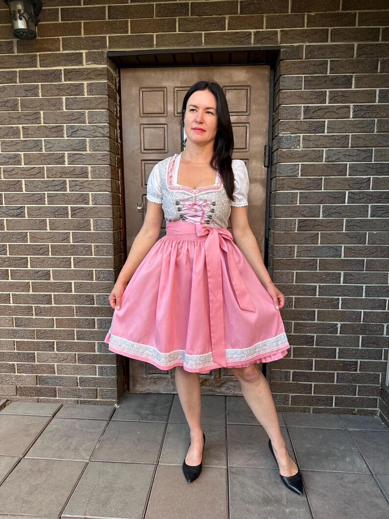 May include: A dirndl dress featuring a white blouse with short puff sleeves, a grey bodice with pink lace-up detailing and metal hooks, and a solid pink skirt with a wide matching waist tie and a decorative white lace trim along the hemline.
