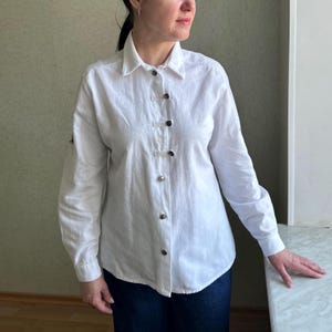 May include: White button-down shirt with long sleeves. The shirt features a classic collar and a series of decorative buttons down the front. The sleeves have buttoned cuffs. The shirt is made of a lightweight fabric.