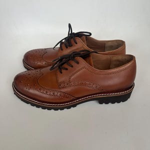 May include: A pair of brown leather Oxford shoes with decorative perforations and black laces. The shoes have a sturdy sole with a white stitched border and a dark brown tread.