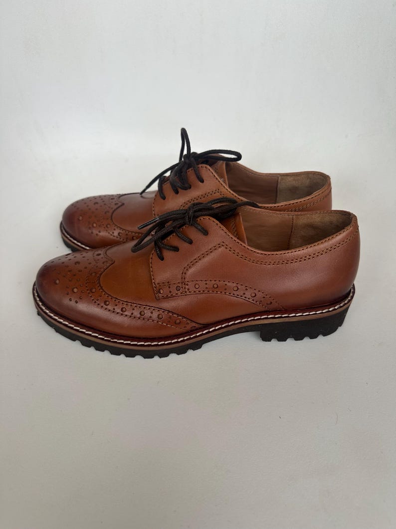 May include: A pair of brown leather Oxford shoes with decorative perforations and black laces. The shoes have a sturdy sole with a white stitched border and a dark brown tread.