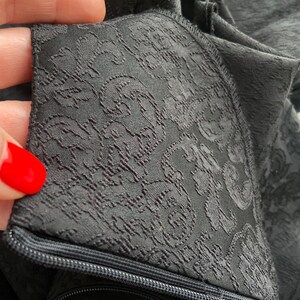 May include: Close-up of a black fabric with a repeating floral pattern. The fabric has a zipper along the bottom edge.