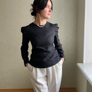 May include: A black long-sleeved blouse with a textured pattern and puffed sleeves. The blouse is paired with white high-waisted pants.