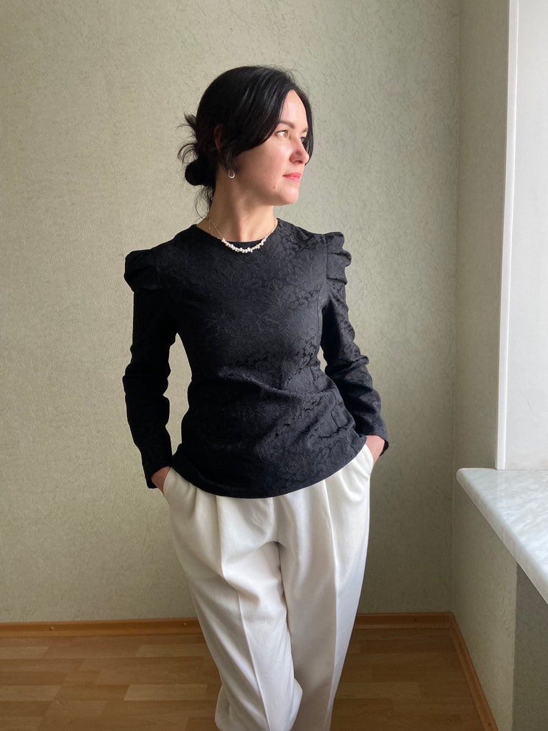 May include: A black long-sleeved blouse with a textured pattern and puffed sleeves. The blouse is paired with white high-waisted pants.
