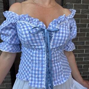 Puede incluir: Una blusa de hombros descubiertos con un patr&oacute;n de cuadros vichy en azul claro y blanco con peque&ntilde;os detalles florales. La prenda incluye mangas cortas abullonadas, ribetes de encaje blanco en el escote y un cord&oacute;n central de cinta azul.