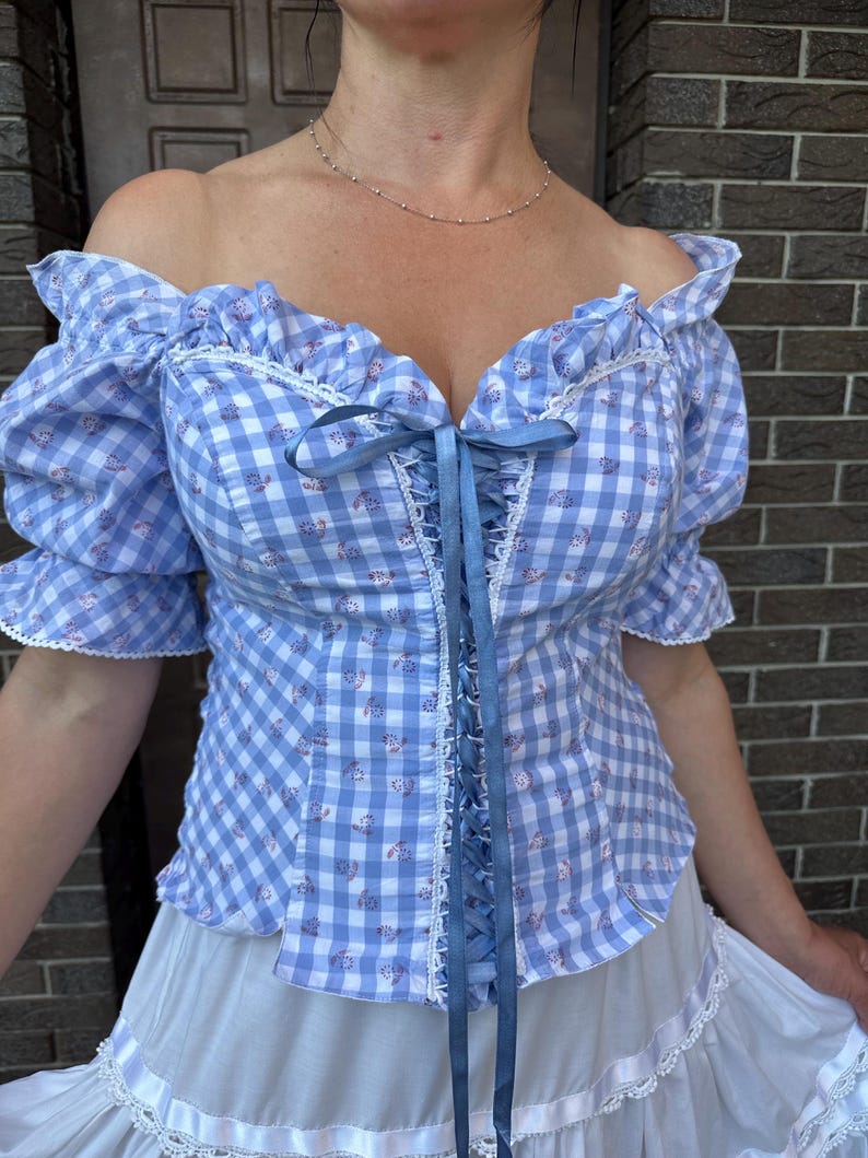 Puede incluir: Una blusa de hombros descubiertos con un patr&oacute;n de cuadros vichy en azul claro y blanco con peque&ntilde;os detalles florales. La prenda incluye mangas cortas abullonadas, ribetes de encaje blanco en el escote y un cord&oacute;n central de cinta azul.