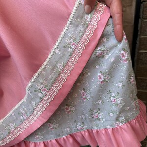May include: A layered skirt featuring a solid pink fabric panel over a grey fabric panel with a floral rose pattern. The garment is accented with white lace trim along the edges and finished with a gathered pink ruffle at the bottom hemline.