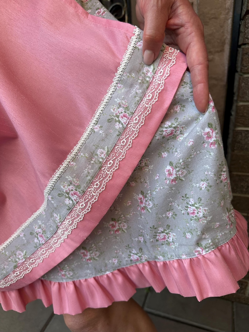 May include: A layered skirt featuring a solid pink fabric panel over a grey fabric panel with a floral rose pattern. The garment is accented with white lace trim along the edges and finished with a gathered pink ruffle at the bottom hemline.