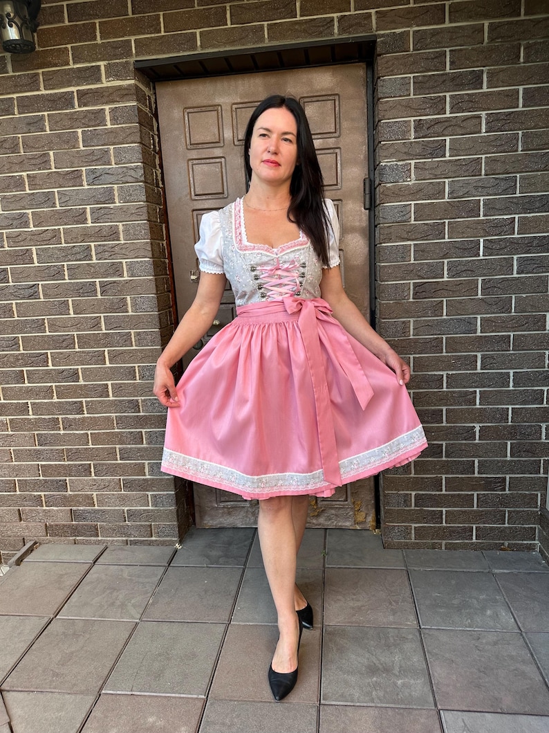 May include: A dirndl dress featuring a white blouse with short puff sleeves, a light gray bodice with a floral pattern and pink lace-up detailing, and a solid pink skirt with a matching floral patterned hem border and a tied pink waist apron.