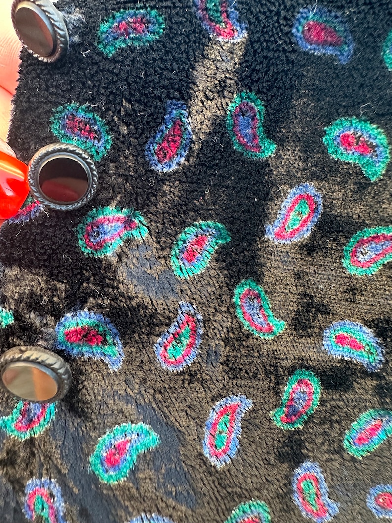 May include: Black velvet fabric with a paisley pattern in shades of blue, green, and red. The fabric has three black buttons with silver accents.