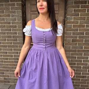 Puede incluir: Un vestido dirndl morado con escote cuadrado y corpiño abotonado. El vestido presenta un estampado de lunares y una falda amplia. Mangas abullonadas blancas asoman por debajo de los tirantes del vestido. El vestido es de estilo tradicional.