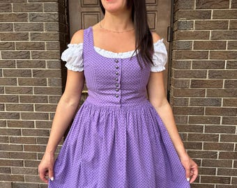 May include: A purple dirndl dress with a square neckline and a button-down bodice. The dress features a polka dot pattern and a full skirt. White puffed sleeves peek out from under the dress's straps. The dress is a traditional style.