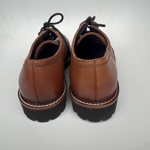 May include: A pair of brown leather shoes with black laces. The shoes have a rounded toe and a thick, black sole with white stitching around the edge. The shoes are shown from the back.