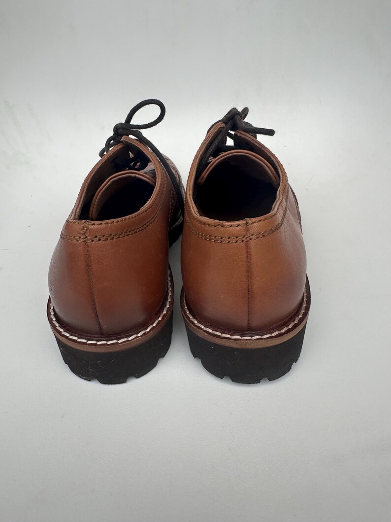 May include: A pair of brown leather shoes with black laces. The shoes have a rounded toe and a thick, black sole with white stitching around the edge. The shoes are shown from the back.