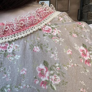 May include: A beige garment featuring a floral pattern with pink roses and green leaves. The neckline is accented with a pink fabric trim, a layer of intricate white lace, and a decorative white braided cord trim along the collar edge.