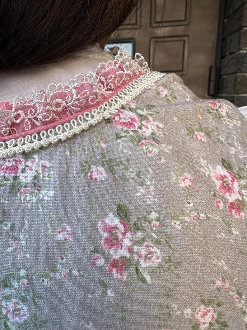May include: A beige garment featuring a floral pattern with pink roses and green leaves. The neckline is accented with a pink fabric trim, a layer of intricate white lace, and a decorative white braided cord trim along the collar edge.