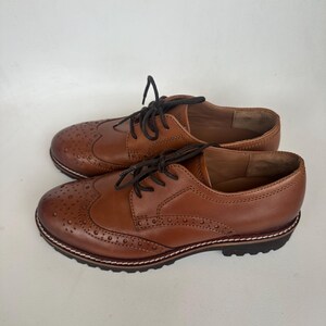 May include: A pair of brown leather Oxford shoes with decorative perforations and dark brown laces. The shoes have a classic wingtip design and a stitched sole. The shoes are a warm brown color.