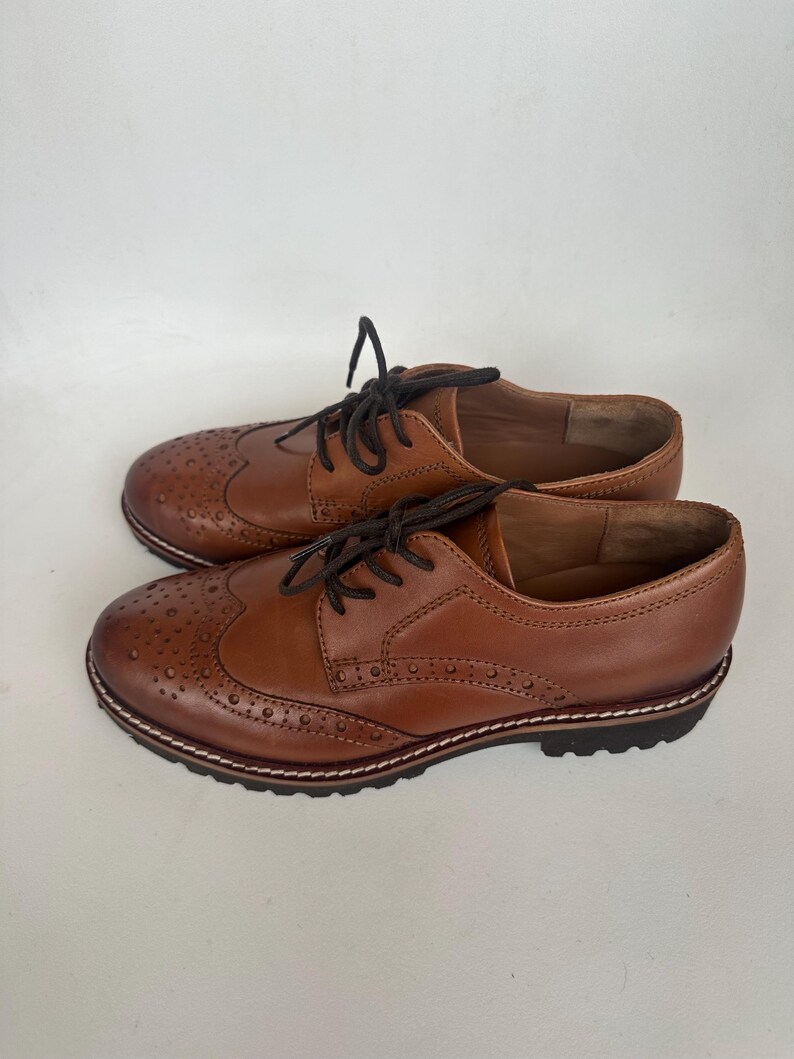 May include: A pair of brown leather Oxford shoes with decorative perforations and dark brown laces. The shoes have a classic wingtip design and a stitched sole. The shoes are a warm brown color.
