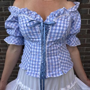 Puede incluir: Una blusa de hombros descubiertos con un patr&oacute;n de cuadros vichy en azul claro y blanco con peque&ntilde;os detalles florales. La prenda incluye mangas cortas abullonadas, escote con ribete de encaje y cierre frontal con lazo azul.