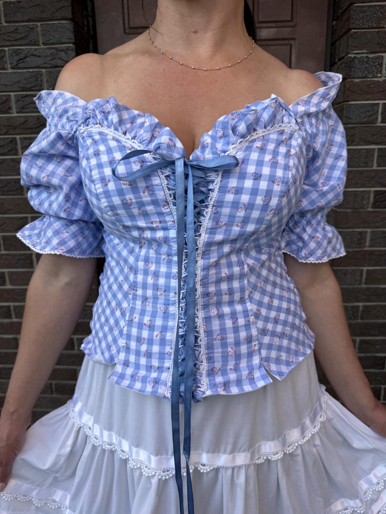 Puede incluir: Una blusa de hombros descubiertos con un patr&oacute;n de cuadros vichy en azul claro y blanco con peque&ntilde;os detalles florales. La prenda incluye mangas cortas abullonadas, escote con ribete de encaje y cierre frontal con lazo azul.