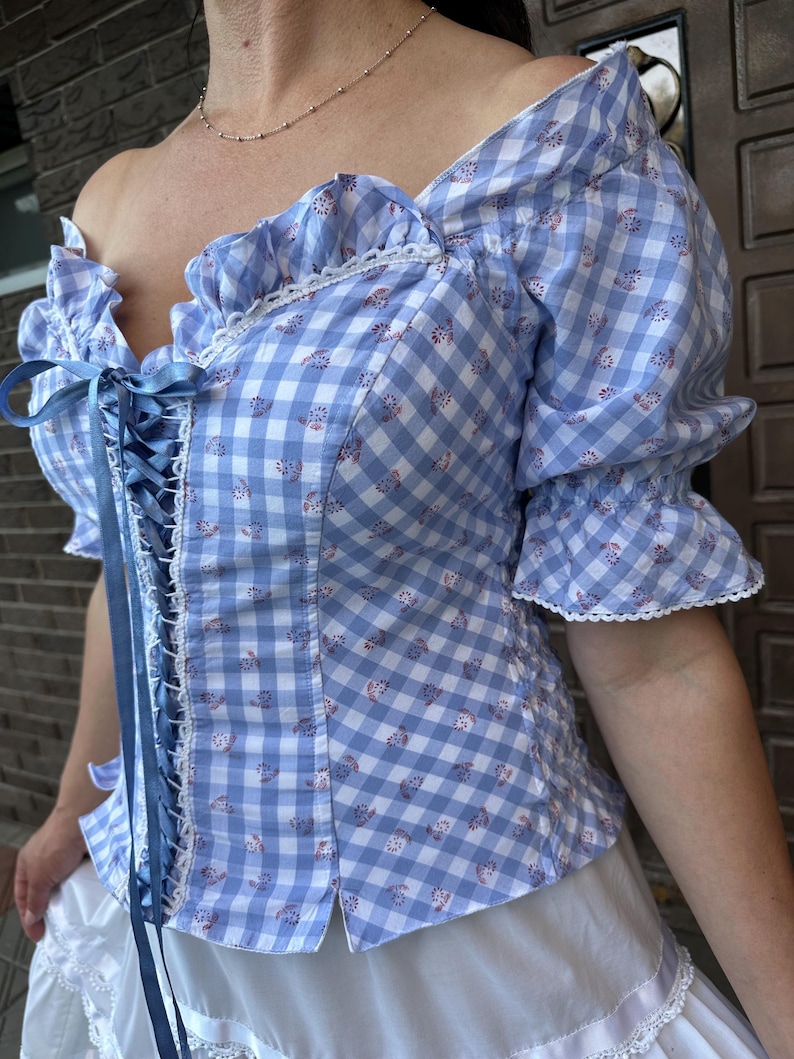 Puede incluir: Una blusa de cuadros vichy en azul claro y blanco con escote coraz&oacute;n, ribetes de volantes y mangas cortas abullonadas. La parte delantera incluye un cierre de cordones con cinta azul, detalles de encaje blanco y un peque&ntilde;o estampado floral.