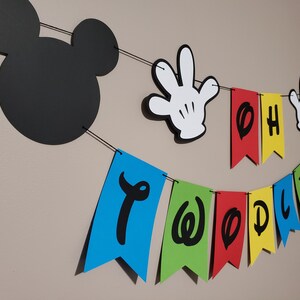 May include: A two-year-old birthday banner with a Mickey Mouse theme. The banner includes a black Mickey Mouse head silhouette, white gloved hands, and colourful pennants spelling "Oh Two Doodles". The pennants are blue, green, red, yellow, and red.