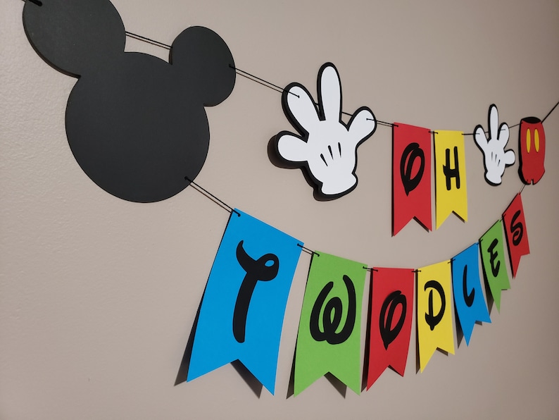 May include: A two-year-old birthday banner with a Mickey Mouse theme. The banner includes a black Mickey Mouse head silhouette, white gloved hands, and colourful pennants spelling "Oh Two Doodles". The pennants are blue, green, red, yellow, and red.