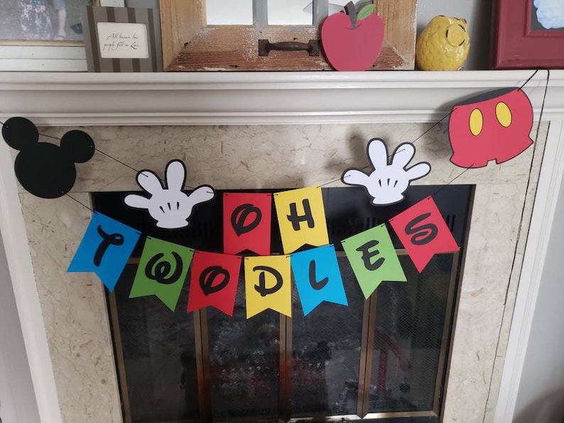 May include: A vibrant banner spelling "TWOODLES" in black letters on individual flags. The banner is decorated with Mickey Mouse-themed cutouts, including a black silhouette of Mickey's head, white gloves, and red shorts.