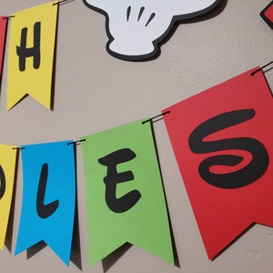 May include: A colourful banner spelling "OH TODDLES" in black letters on red, yellow, blue, and green pennant flags. A white Mickey Mouse hand is visible above the banner. The banner is strung on a black string.