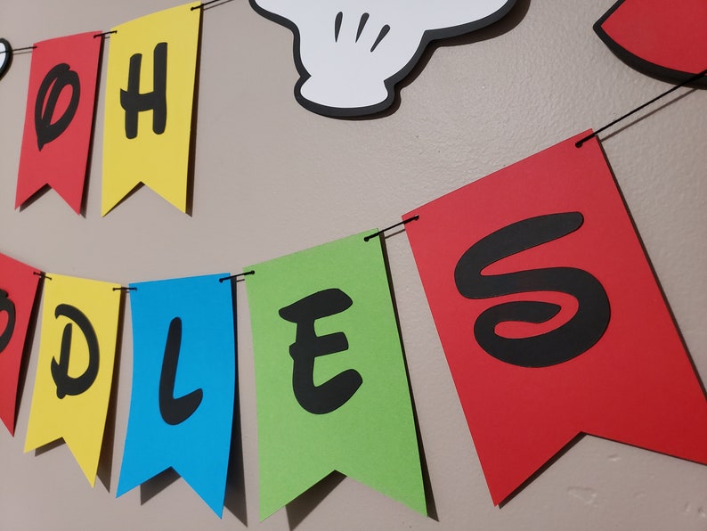 May include: A colourful banner spelling "OH TODDLES" in black letters on red, yellow, blue, and green pennant flags. A white Mickey Mouse hand is visible above the banner. The banner is strung on a black string.