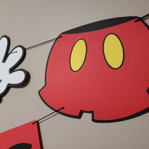 May include: A red, black, and yellow Mickey Mouse-themed banner with a white glove and a red shorts cutout. The banner is strung together with black string, ideal for a birthday party or themed event.