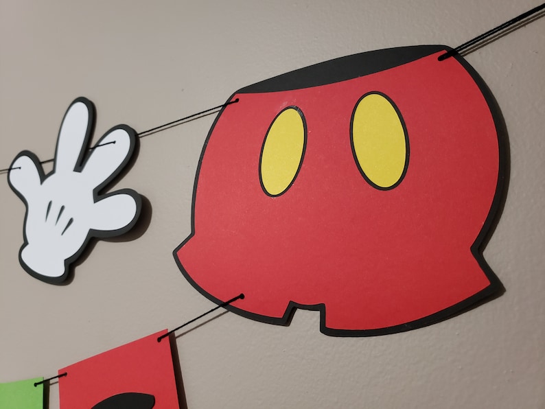 May include: A red, black, and yellow Mickey Mouse-themed banner with a white glove and a red shorts cutout. The banner is strung together with black string, ideal for a birthday party or themed event.