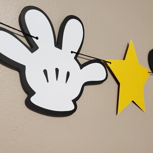 Puede incluir: Una mano de Mickey Mouse en blanco y negro, una estrella amarilla y una cabeza de Mickey Mouse negra est&aacute;n enhebradas en una cuerda negra. La cuerda est&aacute; colgada en una pared.